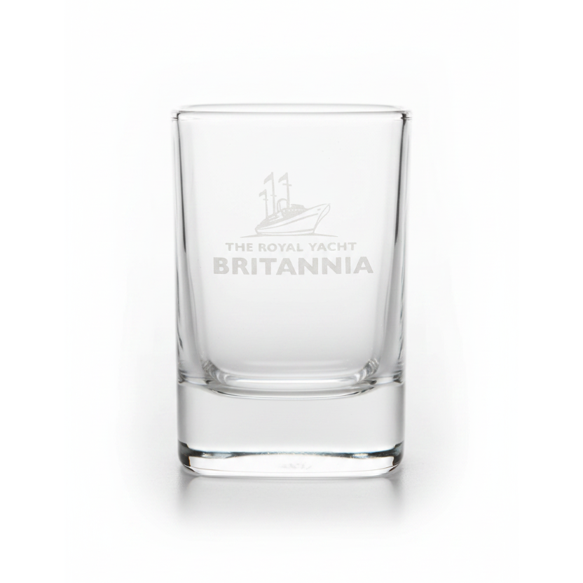 A clear glass shot glass with a white etched logo of a yacht and the text 'THE ROYAL YACHT BRITANNIA' on the front, set against a plain white background.