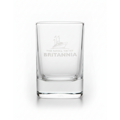 A clear glass shot glass with a white etched logo of a yacht and the text 'THE ROYAL YACHT BRITANNIA' on the front, set against a plain white background.