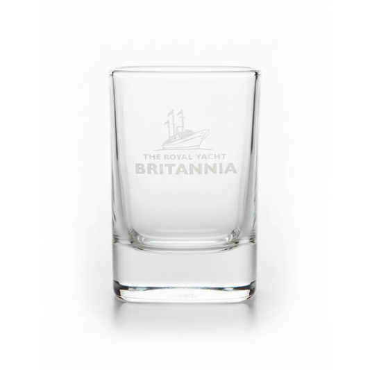 A clear glass shot glass with a white etched logo of a yacht and the text 'THE ROYAL YACHT BRITANNIA' on the front, set against a plain white background.