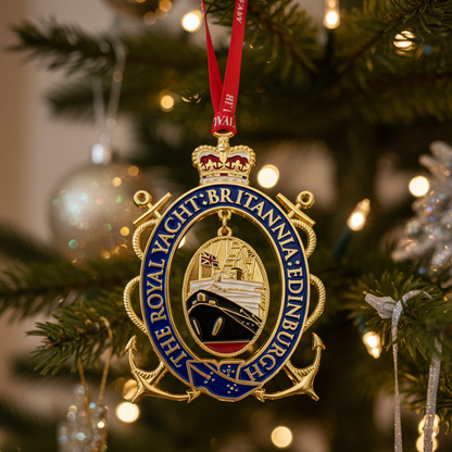 A Royal Yacht Britannia-themed Christmas ornament hanging from a decorated Christmas tree with warm lights and silver ornaments in the background.