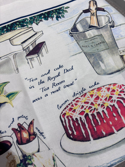A close-up of a printed textile or fabric panel featuring a vintage-style tea and cake illustration. The design includes a red glazed cake with white drizzle, a silver champagne bucket with a bottle, a white piano, and handwritten text reading, 'Tea and cake in the Royal Deck Tea Room was a real treat.